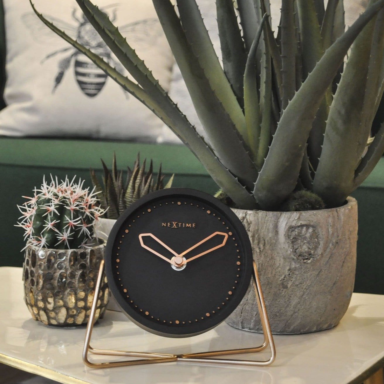 NeXtime Cross Table clock with a black dial and brass stand beside potted plants