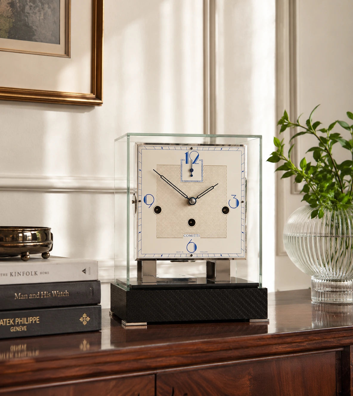 Comitti glass table clock on a polished wood console beside books and flowers